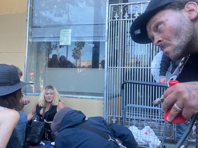 A group of people sit and gather on a sidewalk in front of a building and metal gate; one man in a cap and tattoos is in the foreground holding a red lighter and pipe.