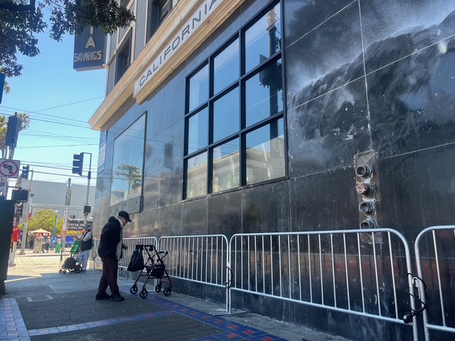 An elderly person with a walker stands on a city sidewalk near a building with black tiles, metal barriers, and reflective windows.