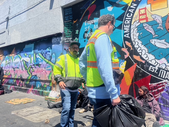 Two workers in safety vests stand near several seated people on a city sidewalk beside a colorful mural and graffiti-covered wall.