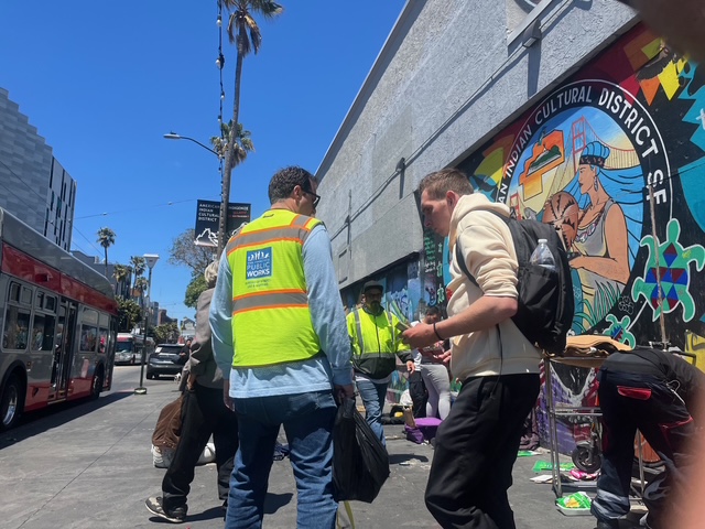 People wearing safety vests interact with others on a city sidewalk near a colorful mural and a parked bus under a clear sky.