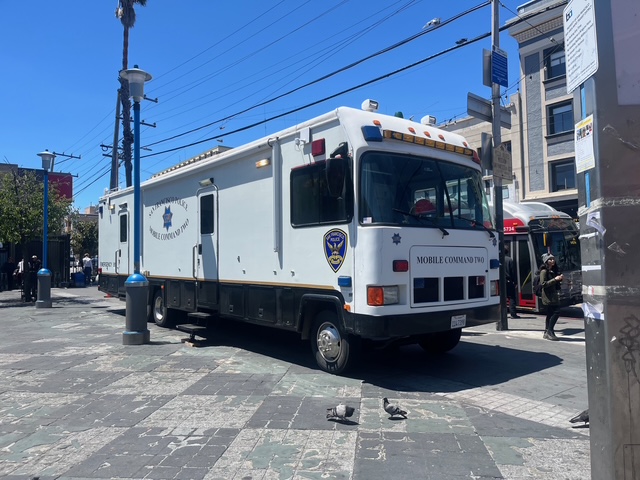 A large white police mobile command unit is parked on a city street near a bus stop, with a few people and pigeons nearby.