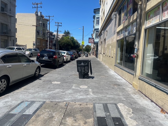 A black trash bin sits in the middle of a sunny city sidewalk lined with parked cars and buildings.
