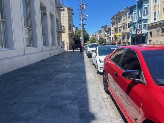 A red car and several other vehicles are parked along a city street lined with multi-story buildings under a clear blue sky.