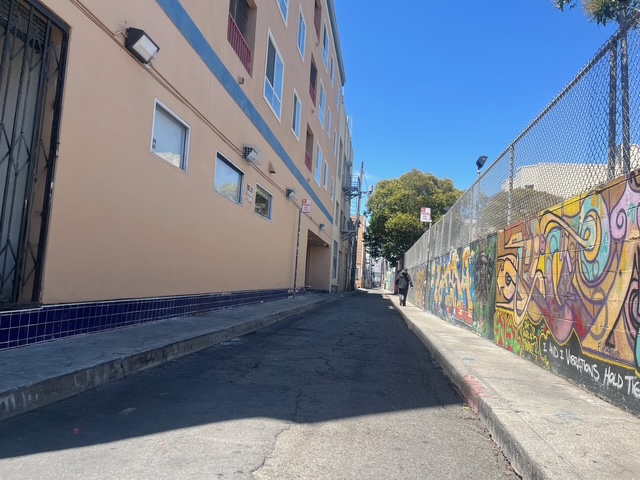 A narrow urban alley with a beige building on the left and a graffiti-covered fence on the right, under a clear blue sky.