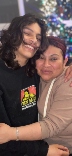 Two people smiling and hugging, standing in front of a blurred background with colorful lights.