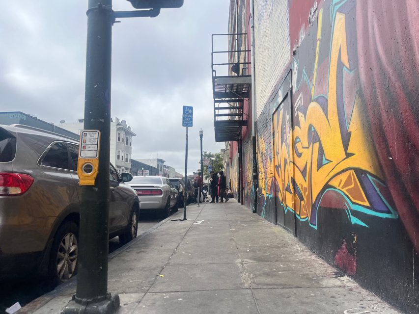 Graffiti covers a wall beside a city sidewalk lined with parked cars outside Yvonne’s, a yellow pedestrian button, and people standing in the distance under a cloudy sky.
