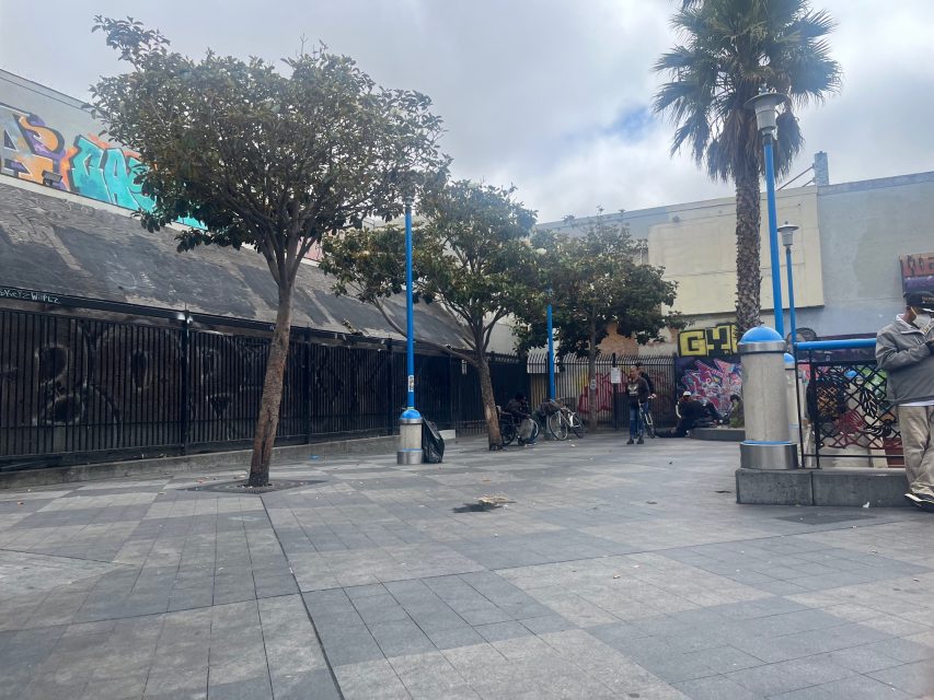 Urban plaza with several trees, graffiti on Yvonne's building walls, scattered trash, and a few people standing and sitting near a metal fence. Sky is overcast.