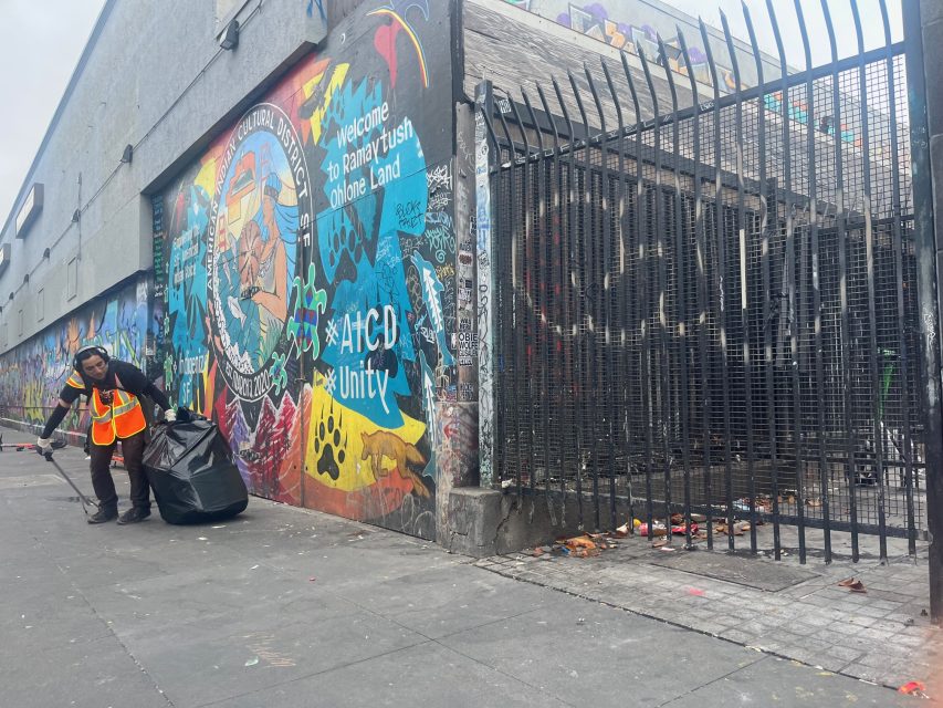 A worker in an orange safety vest collects trash on a city sidewalk in front of Yvonne's colorful mural and a gated area covered with graffiti.