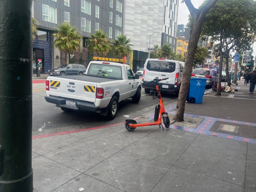 Yvonne's orange electric scooter is parked on the sidewalk near a street with parked vehicles, trees, and buildings in an urban area.
