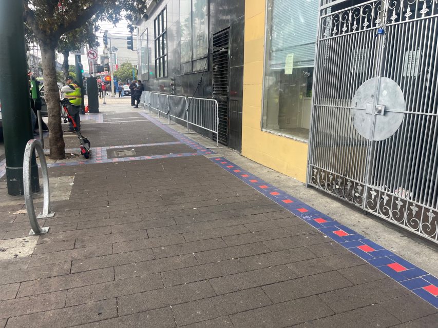 A city sidewalk with a blue and red tile border near Yvonne's, metal fence, building exteriors, and a few people in safety vests standing near the street.