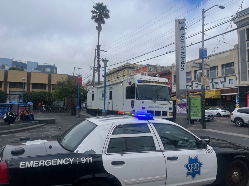 San Francisco police car and Yvonne's mobile command unit parked at 16th Street Mission BART station, with people and buildings in the background.