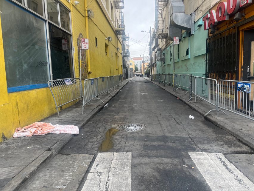 A narrow urban alley with metal barricades on both sides, a yellow building on the left, and Yvonne's taqueria sign on the right. The street is mostly empty and overcast.