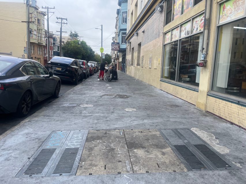 A city sidewalk with parked cars on the left, Yvonne's hotel and restaurant on the right, and people with a stroller and shopping cart in the distance.