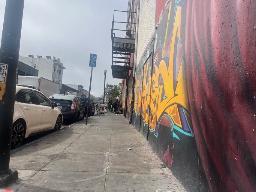 City sidewalk with parked cars on the left, a building with colorful fire-themed graffiti on the right, and a few people gathered in the distance under an overcast sky.