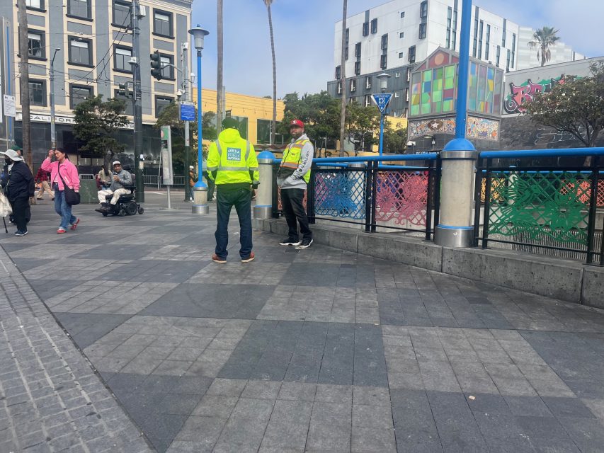 Two people in yellow safety vests stand talking in an urban plaza; pedestrians and a person using a wheelchair are also visible in the background, suggesting coordinated fire safety efforts.