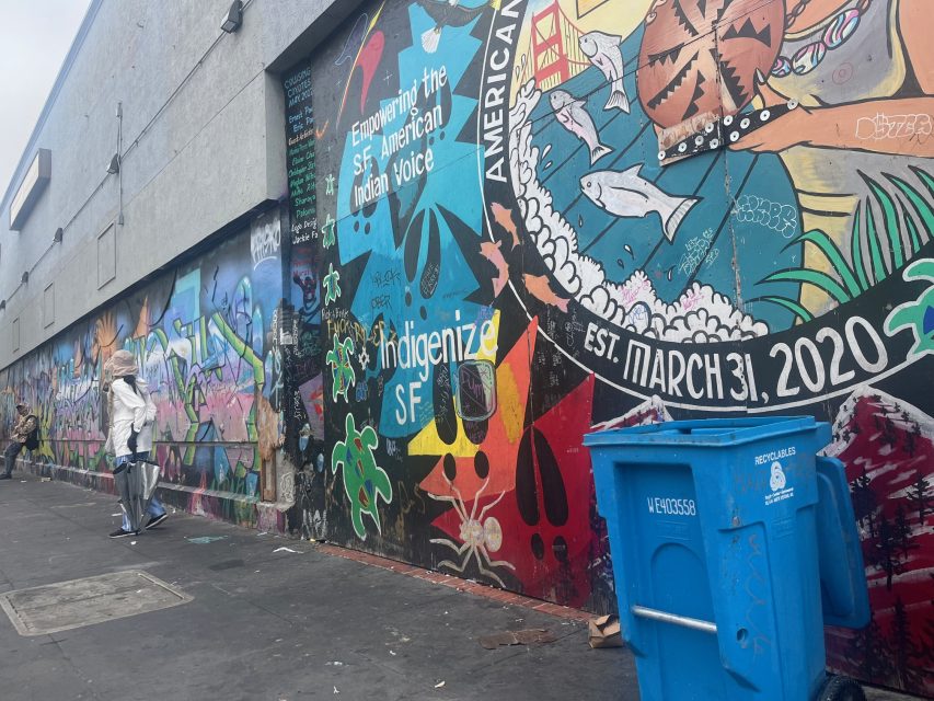A person walks past colorful murals on an urban wall, featuring Indigenous themes of fire and messages, with a blue recycling bin in the foreground.
