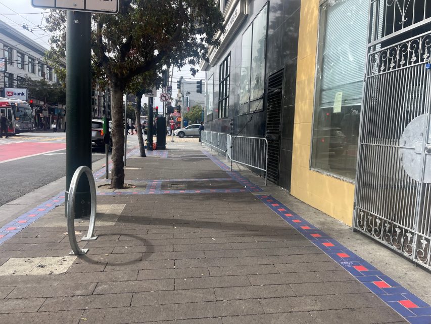 Sidewalk with a blue and red border, bike rack, tree, and metal gate on the right; street with a bus and buildings in the background—a fire hydrant stands nearby for added safety.