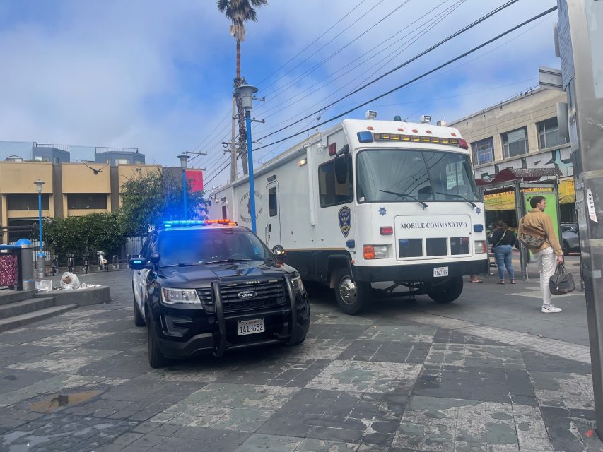 A police car with flashing lights, a mobile command vehicle, and a fire truck are parked in a city square, with people walking nearby and buildings in the background.