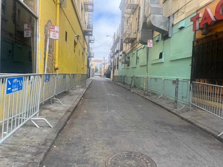 A narrow urban alley lined with metal barricades, yellow and green building walls, and "No Parking" signs, with a fire hydrant on the corner and a partly cloudy sky above.