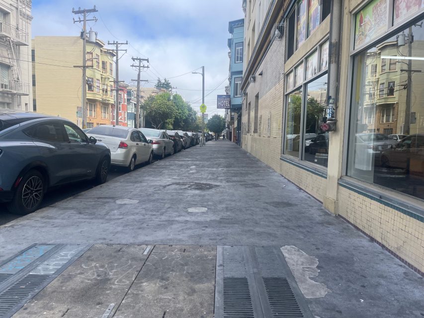 A city sidewalk lined with parked cars on one side and lively storefronts on the other, under a partly cloudy sky, catches the warm glow of fire-like sunset hues.