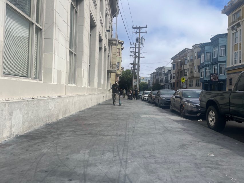 A person rides an electric scooter on a wide urban sidewalk beside a row of parked cars and multi-story buildings, while fire engine lights flash in the distance on a cloudy day.