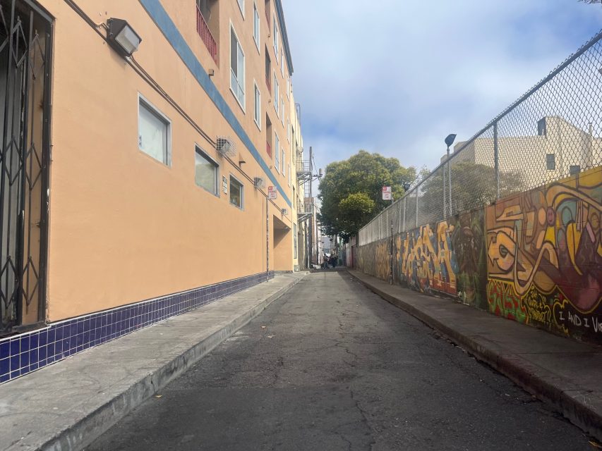 A narrow urban alleyway lined with a peach-colored building on the left and a graffiti-covered wall with a chain-link fence on the right, under a partly cloudy sky, hints of fire-orange paint flaring up amidst the street art.