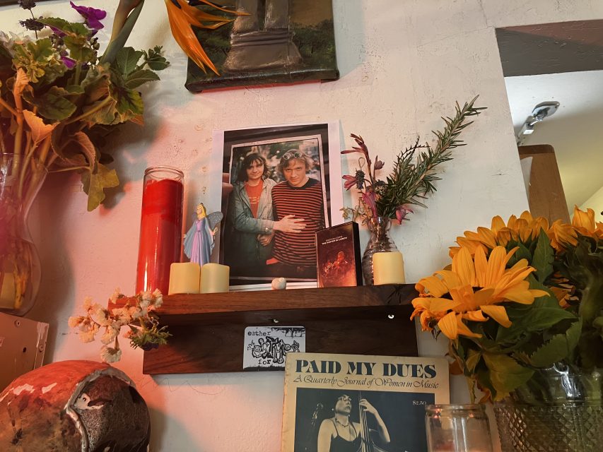A wooden shelf displays a framed photo of two people, candles, a small statue, flowers, a lighter, and a "Paid My Dues" magazine below.
