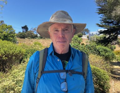 An older man wearing a wide-brimmed hat, blue shirt, and backpack stands outdoors in a sunny, grassy area, watching a butterfly flutter near the bushes and trees in the background.