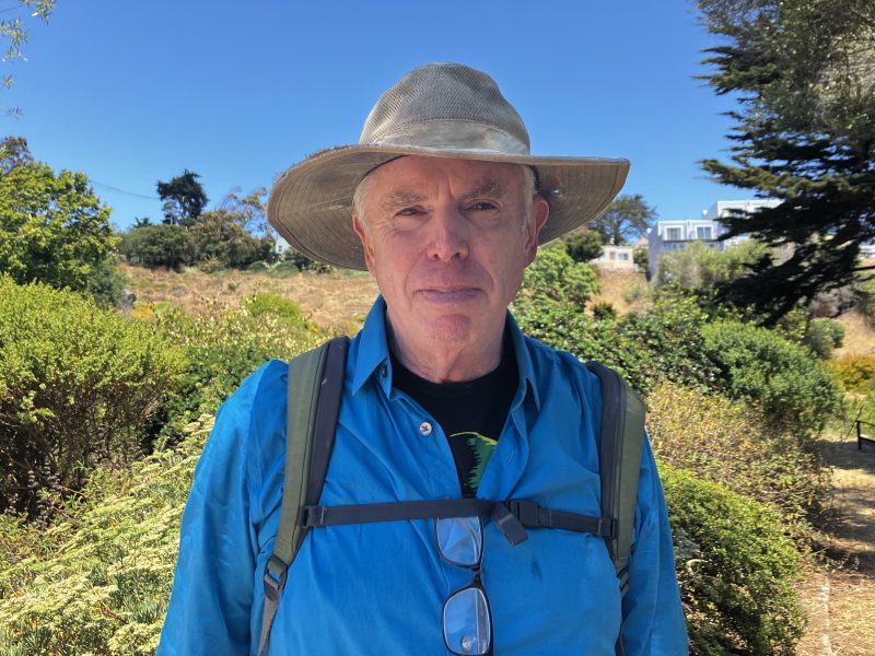 An older man wearing a wide-brimmed hat, blue shirt, and backpack stands outdoors in a sunny, grassy area, watching a butterfly flutter near the bushes and trees in the background.