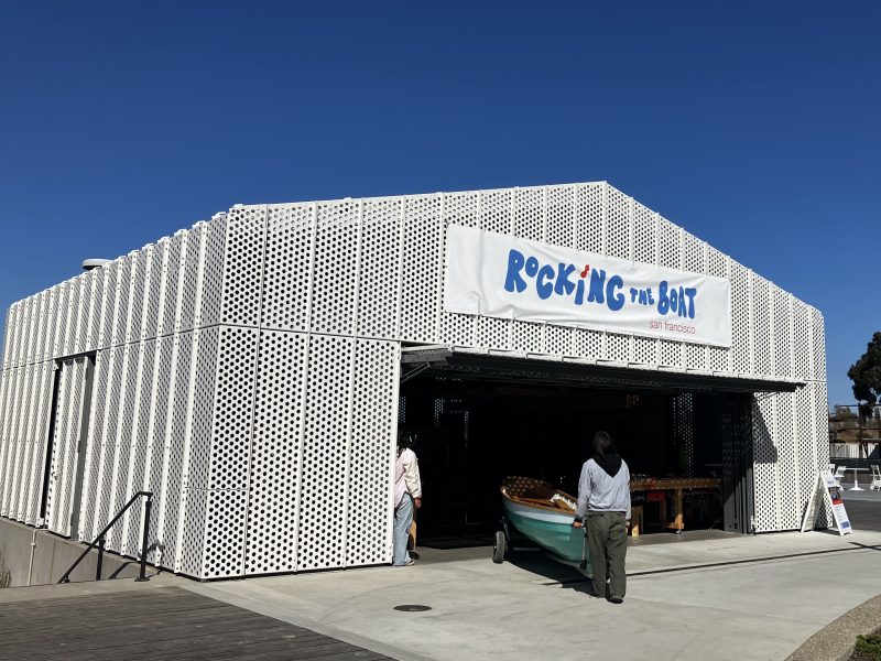 A modern, white perforated metal building features a prominent “Rocking the Boat” banner above the entrance; inside, two people stand near a small wooden boat.