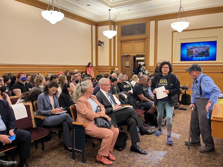A group of people sits in a conference room, some talking and others listening, while two individuals stand and walk near the front.