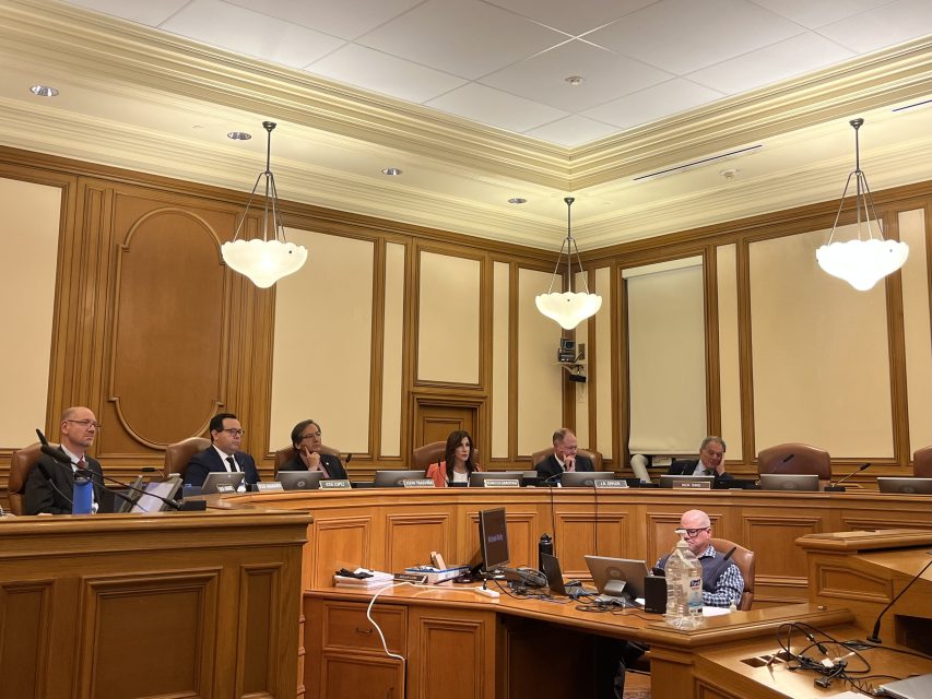 A group of officials sit at a panel in a wood-paneled government meeting room with microphones and laptops on the desks.