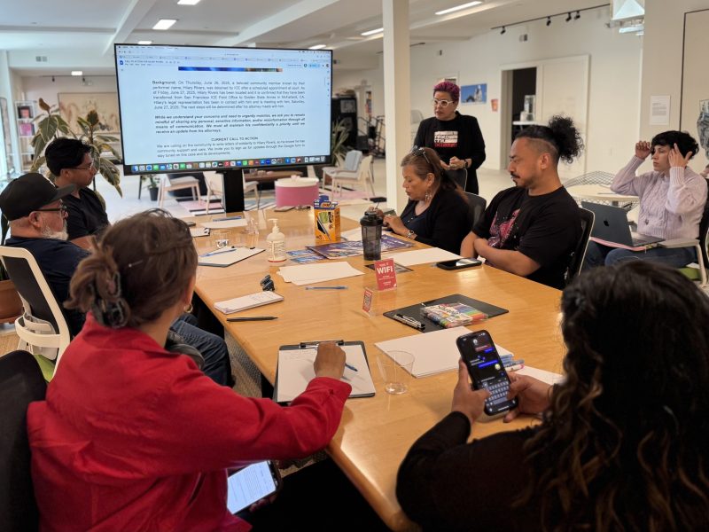 A group of people sit around a table in a meeting room, listening to a presenter standing by a large screen displaying a document.