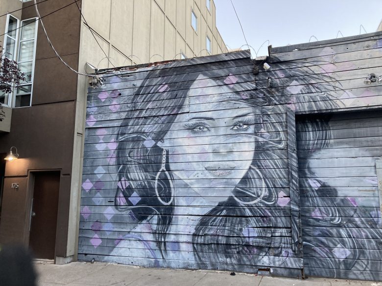 A mural of a woman with long hair and hoop earrings is painted in shades of gray and purple on the side of a wooden building.