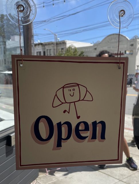 A sign with a smiling croissant cartoon and the word "Open" hangs on a glass door with the street and shops visible outside.