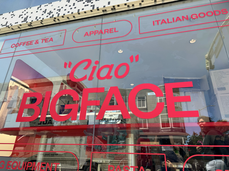 Storefront window with large red text reading "Ciao" and "BIGFACE," along with sections labeled "APPAREL," "COFFEE & TEA," and "ITALIAN GOODS." Reflections of buildings are visible.