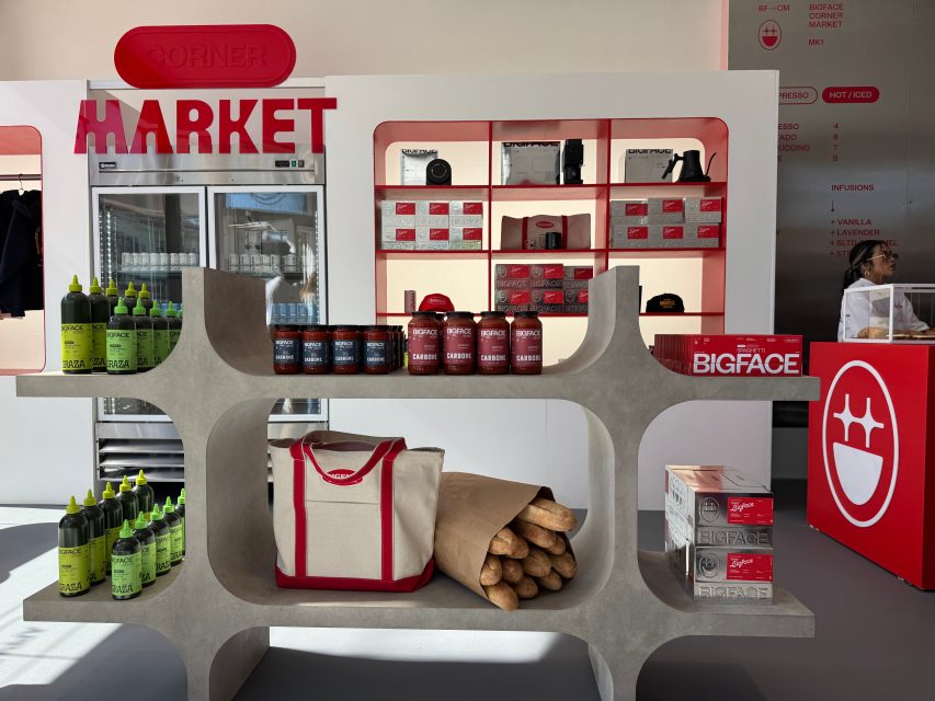 A market display features shelves with bottled drinks, canned goods, a tote bag with baguettes, and boxed items. Refrigerated products and branded bags are visible in the background.