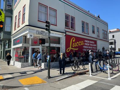 People stand in line outside Lucca deli and market on a sunny day at the corner of 22nd Street and Valencia Street; bicycles are parked nearby.