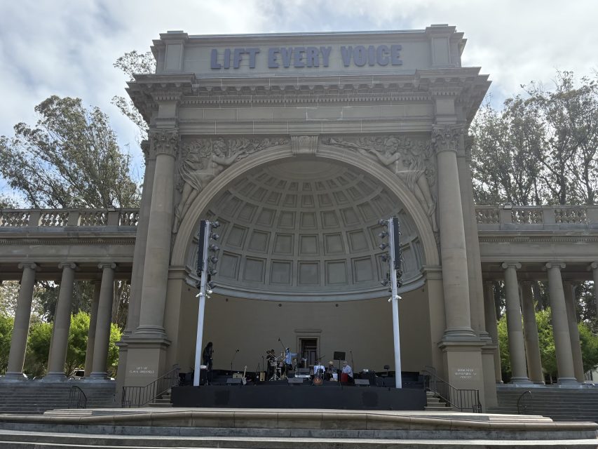 Outdoor band performance at a large open-air bandshell with "LIFT EVERY VOICE" inscribed at the top, surrounded by columns and trees in the background.