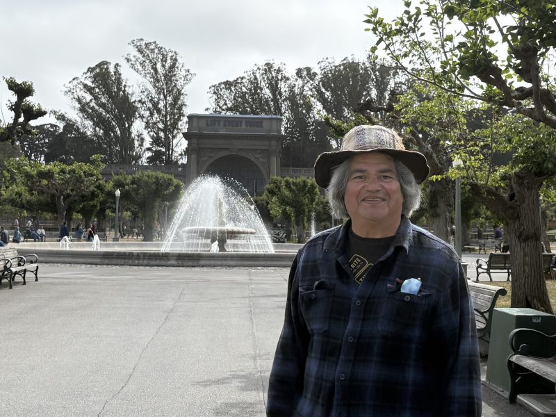 An older man in a plaid shirt and hat stands smiling in front of a large fountain and trees in a park, with a building labeled "Lily Pond House" in the background.
