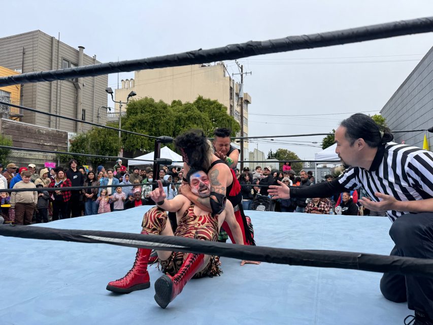Two wrestlers compete in a wrestling ring outdoors as a referee gestures nearby and a crowd watches from behind the ropes.