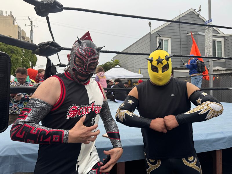 Two masked wrestlers stand posing in front of a wrestling ring outdoors, with people and tents visible in the background.