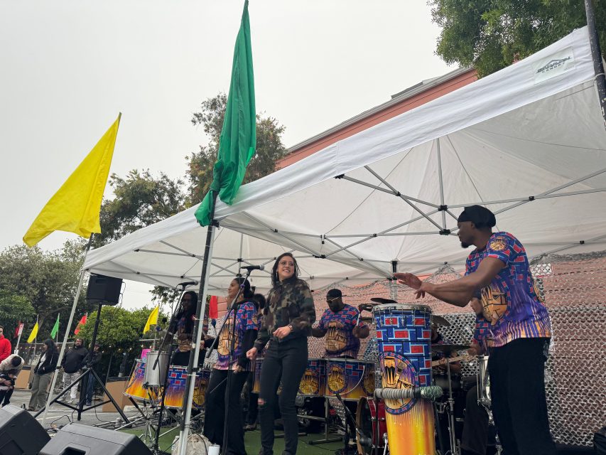 A band dressed in colorful outfits performs on an outdoor stage under a white canopy, accompanied by a woman standing at the front.