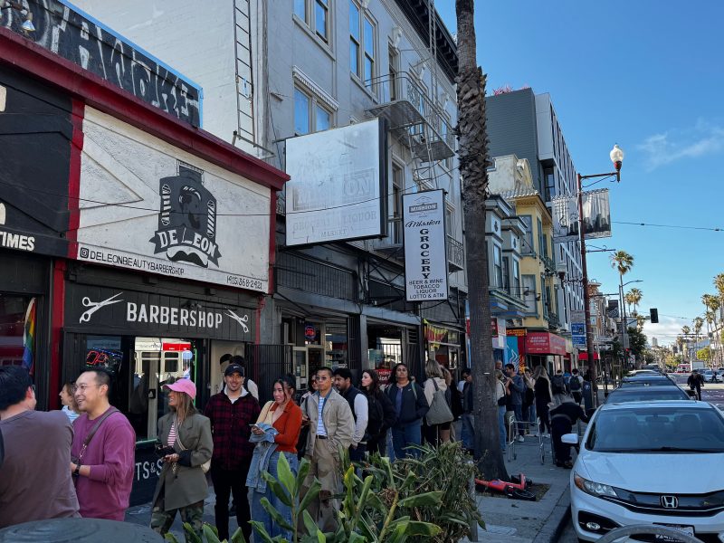 A group of people stands in a line on a city sidewalk outside shops, including a barbershop, on a sunny day with parked cars along the street.