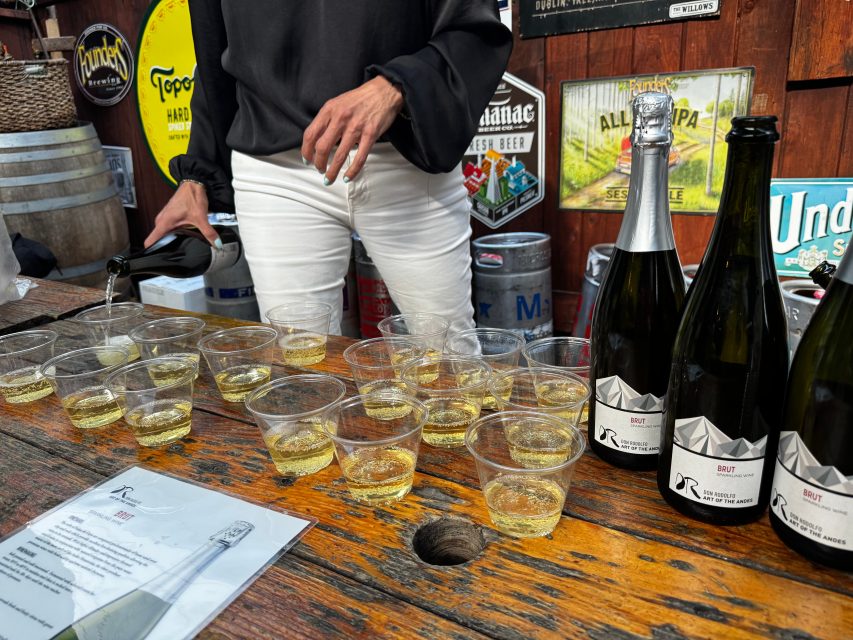 Person in black top and white pants pours sparkling wine into plastic cups on a wooden table, with bottles, flyers, and beer signs in the background.