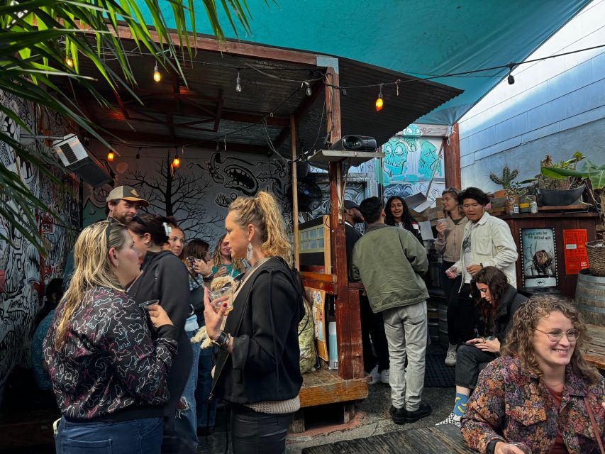 A group of people socializing in an outdoor bar area with graffiti-covered walls, string lights, and plants under a blue tarp.