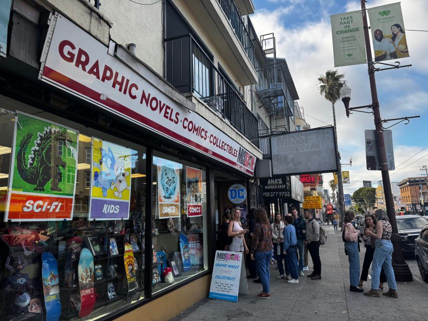 People stand in line outside a comic book store with a sign reading "Graphic Novels · Comics · Collectibles," located on a busy city street with shops and parked cars.