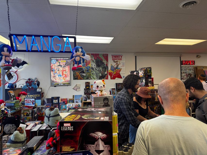 A group of people stand inside a comic and toy store with shelves of collectibles, manga, and a blue neon "MANGA" sign above. Sonic the Hedgehog plush toys hang near the ceiling.