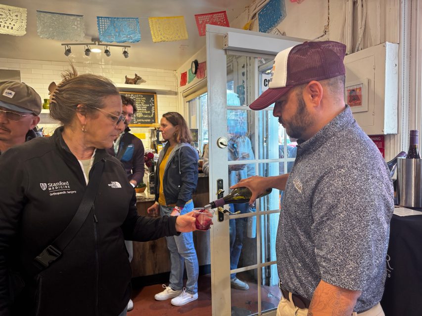 A man pours red wine into a woman's glass inside a casual indoor setting, with other people standing and chatting in the background.