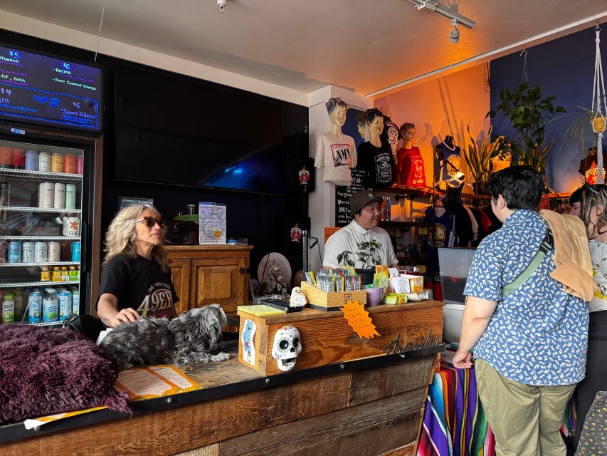 A woman with sunglasses stands behind a counter with a dog, talking to two customers in a colorful shop with shelves, plants, and a refrigerator in the background.